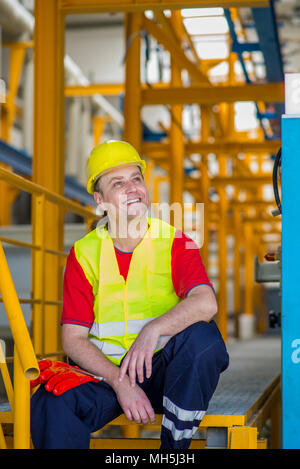 Felice lavoratore in giallo riflettente suit con casco giallo seduto in una fabbrica e sorridente Foto Stock