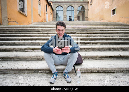 Bel giovane in abbigliamento sportivo con capelli ricci seduti sulle scale e lavorando su tablet Foto Stock