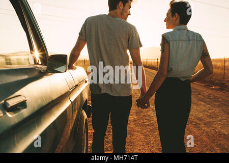 Coppia romantica in piedi tenendo le mani accanto a loro pickup truck durante un viaggio nel paese di lato. Vista posteriore del giovane tenendo le mani e piedi su Foto Stock