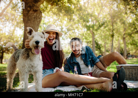 Sorridente coppia giovane su picnic al parco con il loro cane. Ridendo donna accarezza un cane con uomo seduto da nel parco. Foto Stock