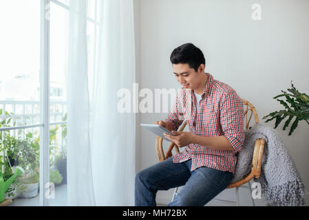 Bel uomo seduto rilassato dalla finestra e utilizzando il tablet Foto Stock