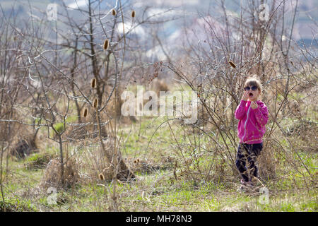 Ritratto di carino piccolo confuso ragazza bionda in rosa casual abbigliamento e occhiali da sole scuri in piedi da solo perso tra secco boccole coccolone e chiedendo per lui Foto Stock