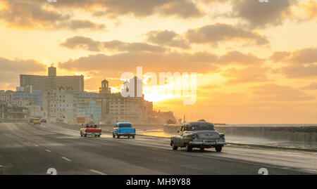 Onde infrangersi come il sole tramonta come classic cars corsa lungo il Malecon a l'Avana, Cuba. Foto Stock