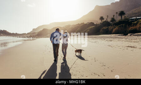 Coppia matura passeggiando lungo la spiaggia con il loro cane. Vista posteriore shot di amorevole coppia matura sulla spiaggia con il cane. Foto Stock