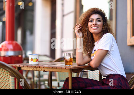 Giovani arabi donna sorridente e seduto in un bar urbano in strada. Ragazza araba in abiti casual di bere una soda all'esterno. Foto Stock