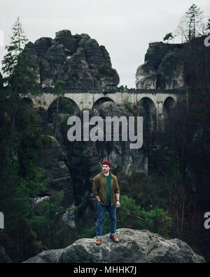 Hipster uomo sulla roccia e guardando a incredibili montagne, foreste e Bastei bridge in Svizzera sassone in autunno, Germania. Foto Stock