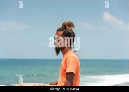 Bandiera Rock, Galle, Sri Lanka: Ritratto di un uomo con lunghi capelli ricci, legato alla moda e una barba, in piedi che guarda al mare. Vista di profilo. Foto Stock