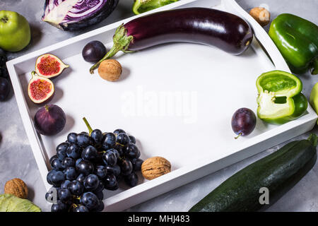 Fresche materie organiche di verde e di colore viola le verdure e la frutta nel vassoio bianco su sfondo di pietra. Copyspace. Foto Stock