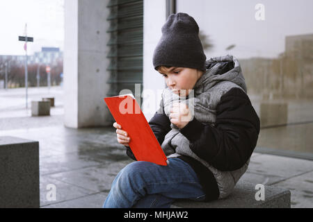 Eccitato ragazzino giocando online gioco sul computer tavoletta mentre seduto fuori da solo sulla strada di città in inverno, celebrando la vittoria con pugno chiuso Foto Stock