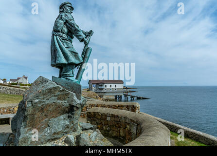 La statua di Richard Evans Lifeboatman si eleva alto con Moelfre scialuppa di salvataggio casa sullo sfondo su Anglesey, Galles del Nord. Foto Stock