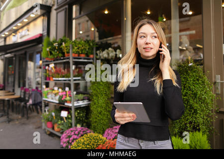 Imprenditrice in piedi con la tavoletta e parlare tramite smartphone vicino al negozio di fiori. Foto Stock