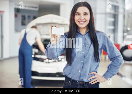 Una bella donna mantiene una business card del car service center. Il meccanico ispeziona la vettura sotto il cofano in background Foto Stock