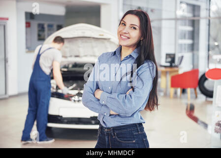 Donna presso un garage auto come ottenere assistenza meccanica. Il meccanico lavora sotto il cofano della macchina Foto Stock