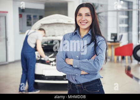 Donna presso un garage auto come ottenere assistenza meccanica. Il meccanico lavora sotto il cofano della macchina Foto Stock