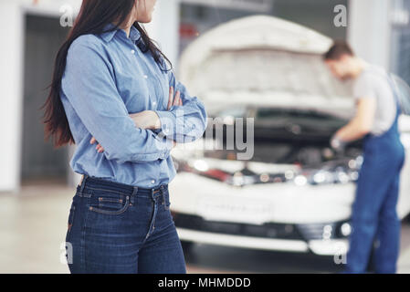 Donna presso un garage auto come ottenere assistenza meccanica. Il meccanico lavora sotto il cofano della macchina Foto Stock