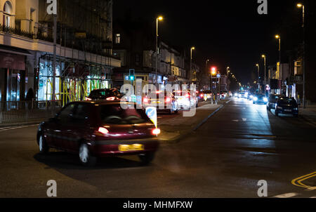 Auto su una strada in una cittadina britannica durante la notte con le luci accese in Inghilterra, Regno Unito. Foto Stock