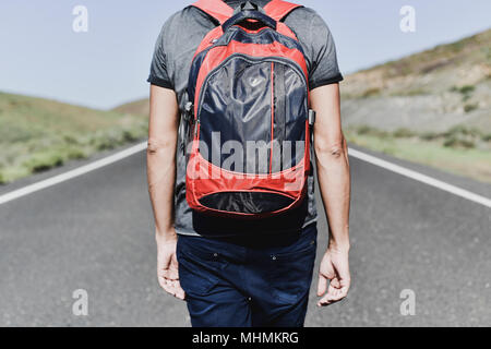 Primo piano di un giovane uomo caucasico, visto da dietro, portando uno zaino camminando lungo una strada secondaria Foto Stock