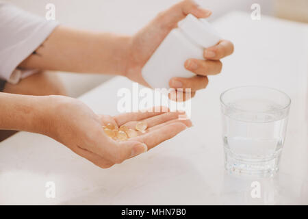 Mano richiede capsule giallo di omega 3, bianco pillole di glucosammina e il calcio su scatola di plastica, vetro con acqua al tavolo di legno, vista dall'alto. Foto Stock