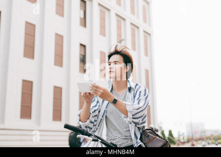Elegante uomo chattare mentre si utilizza il telefono cellulare mentre è seduto sulla bicicletta, all'esterno. Vestito in plaid shirt, t-shirt e jeans. Close-up. Foto Stock