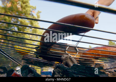 Cani grigliare su una cremagliera su un fuoco aperto nel camino su una sera d'estate. Una persona con barbecue a tenaglia maniglia visibile il grill. Foto Stock