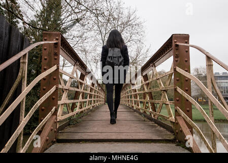 Una giovane donna con i capelli neri e vestito di nero da soli passeggiate attraverso un ponte pedonale in acciaio attraversando un piccolo fiume. Foto Stock