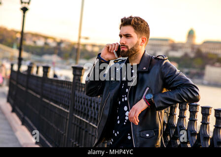 Un bel giovane hipster uomo appoggiato alla recinzione al tramonto mentre si parla sul suo smartphone Foto Stock