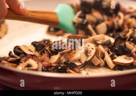 Mano con spatola per mescolare un trito di olive nere, le cipolle rosse e funghi tagliati a fettine in una padella. Foto Stock