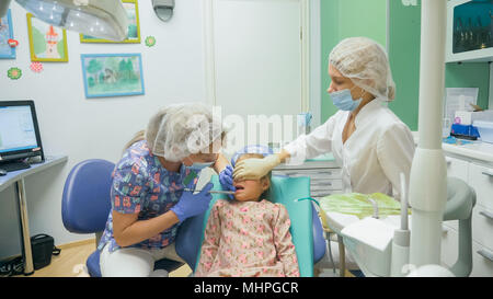 Bambino con una madre a un dentista di ricezione. La ragazza si trova in cattedra, dietro la madre. Il dottore lavora con un assistente. Procedura per la perforazione di un dente. Impostazione di una sigillatura temporanea. Foto Stock