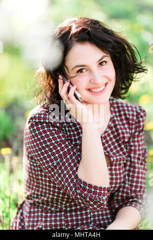 Business, femminilità, concetto di contatto. vi è bella donna nel vestire con piccoli fiori rossi, lei sta ascoltando qualcuno dal suo telefono e sorridente felicemente, i suoi occhi scuri sono incandescente Foto Stock