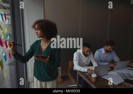 Architetto femmina con tavoletta digitale la scrittura su una nota adesiva mentre i colleghi utilizzando il computer portatile Foto Stock