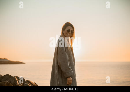 Ritratto di donna che indossa gli occhiali da sole e cappotto invernale guardando la telecamera sorridendo, vista laterale, Odessa, Odeska, Oblast di Ucraina Foto Stock