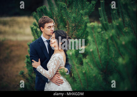 Appena sposata giovane teneramente bacio tra abeti Foto Stock