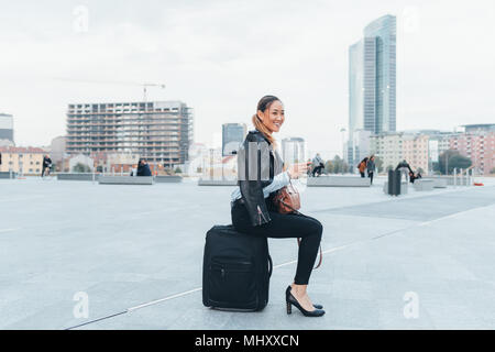Ritratto di imprenditrice all'aperto, seduto sulla valigia, sorridente Foto Stock