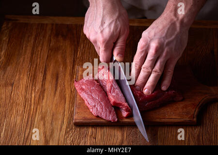 Preparazione per la cena. La cottura, la lavorazione delle carni. Strong professional mani dell'uomo taglio bistecca di manzo crudo, il fuoco selettivo di close-up. Sicuro di sé Foto Stock