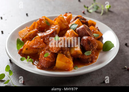 Patate spezzatino di maiale con le fette di salsiccia e salsa di pomodoro su una piastra bianca con origano in foglie Foto Stock