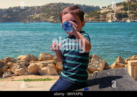 Sete piccolo ragazzo seduto su una panca di legno e di bere acqua minerale. Sullo sfondo di una porta del Mediterraneo. Foto Stock