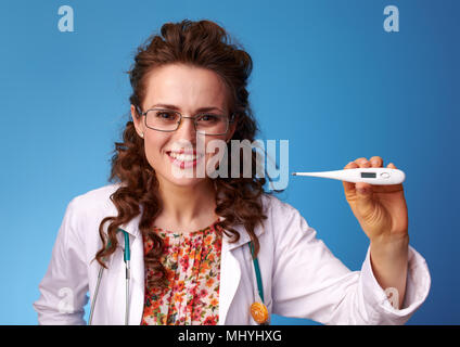 Sorridente medico pediatra in bianco manto medico mostrando termometro contro sfondo blu Foto Stock