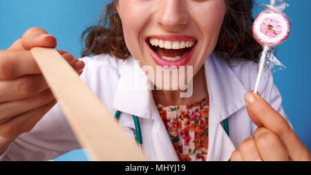 Felice paediatrist medico in bianco manto medica con lollipop usando una spatola per esaminare gola isolato su blu Foto Stock