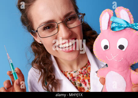 Sorridente paediatrist medico in bianco manto medica con la siringa a distrarre il bambino con il giocattolo isolato su sfondo blu Foto Stock