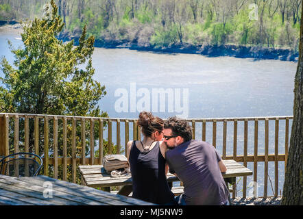 Rocheport, Missouri, Stati Uniti d'America, 30 Aprile 2018: Coppia giovane seduto lungo il fiume; uomo teneramente baciando la alle spalle della donna; fiume e boschi nel backgro Foto Stock
