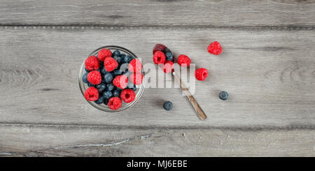 Tabella vista dall'alto sulla piccola ciotola di vetro riempito con un mix di mirtilli e lamponi, cucchiaio d'argento sul lato, posto su grigio scrivania di legno. Foto Stock