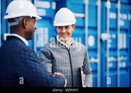 Due ingegneri sorridente indossando hardhats agitando mani insieme mentre in piedi da contenitori di merci in un cantiere di spedizione Foto Stock