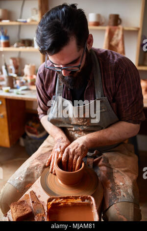 Ritratto di un bel maschio barbuto potter in plaid shirt e grembiule sporco che stampi ciotola da argilla, il fuoco selettivo di close-up. Il lavoro creativo processo. Foto Stock