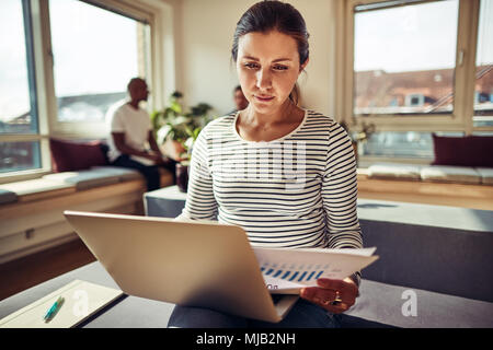 Focalizzato giovane imprenditrice lavorando su un computer portatile e la lettura dei documenti mentre è seduto sulla sua scrivania in ufficio con i colleghi parlando in background Foto Stock