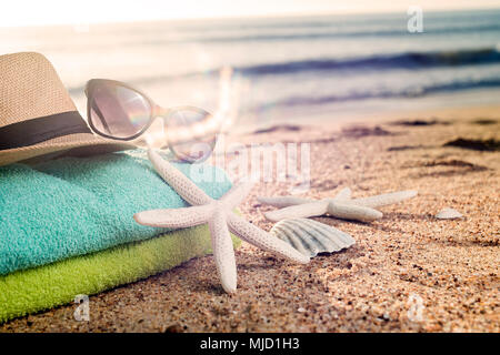 Estate accessori come asciugamani colorati, cappello di paglia, occhiali da sole e conchiglie sulla spiaggia sabbiosa Foto Stock