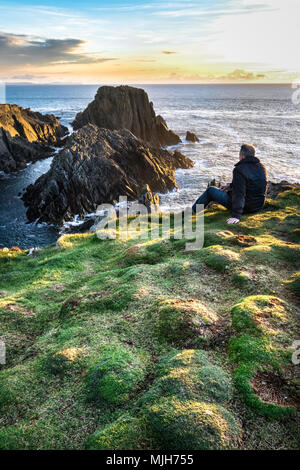 Un uomo seduto sul bordo di una scogliera sul mare che guarda all'oceano al tramonto. Questa foto è stata scattata a Malin Head Irlanda Foto Stock