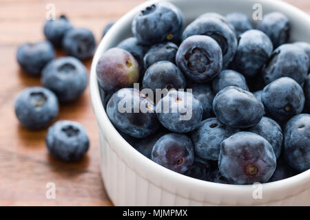 Di piccole dimensioni e di colore bianco ciotola riempita con mirtilli freschi, sei mirtilli sulla sinistra, tavola di legno sfondo, mangiare sano concetto Foto Stock