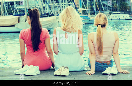 Tre giovani ragazze seduti sul pontile in legno e guardando su barche in marina europea Foto Stock