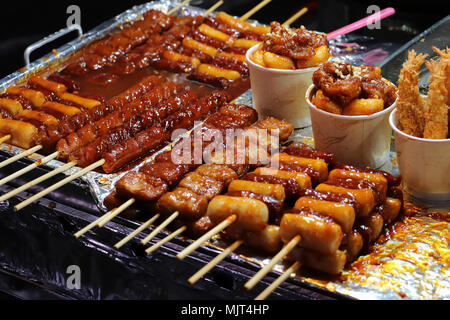 Close-up di salsicce e carne su spiedini di legno ricoperta con una salsa rossa, la cottura su una griglia su una strada posteriore a Seul, in Corea del Sud di notte. Foto Stock