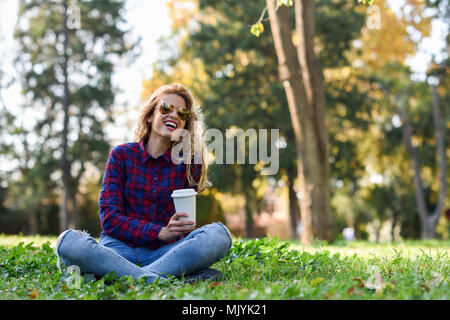 Giovane donna felice in camicia a scacchi e jeans blu con sorriso toothy e occhiali da sole. Ragazza bionda di bere il caffè nel parco seduto sull'erba indossando cas Foto Stock